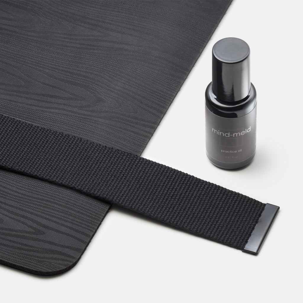 close up of the rounded edge of a wood grained textured yoga mat in a dark gray with black woven polyester fabric yoga strap and black mind-meld practice oil bottle visible against a white background.