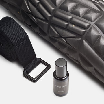 closeup of black foam roller with embossed surface design, a riolled up black yoga strap and mind-meld practice oil bottle visible.